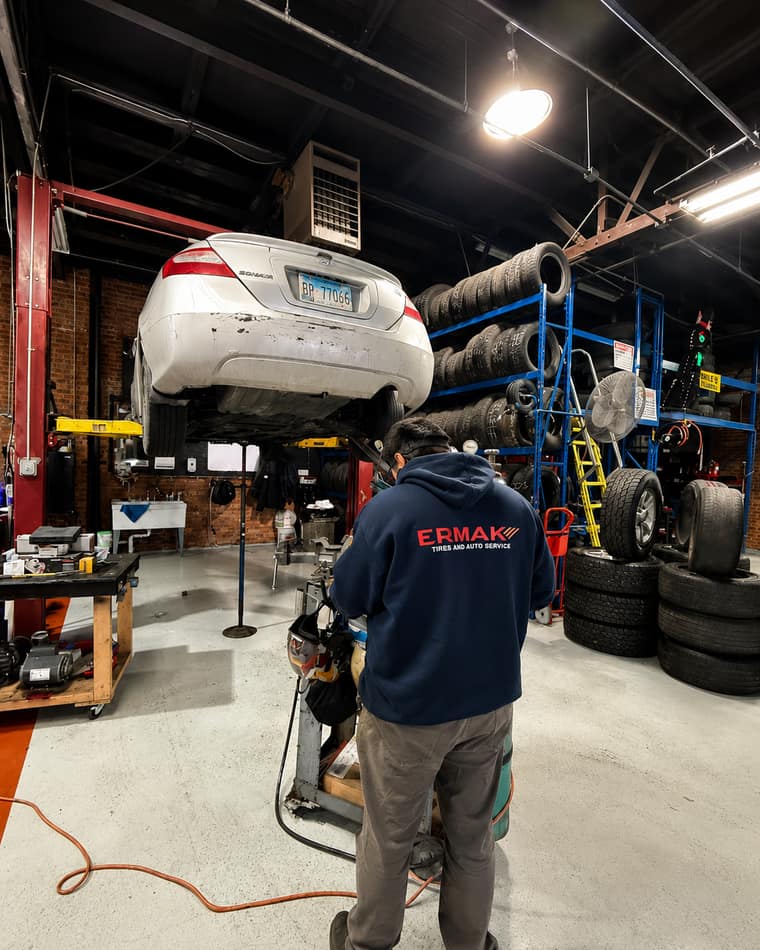 An ERMAK Tires technician in a branded hoodie working under a vehicle in the service bay.